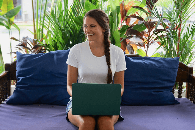 Widget Boxes Nicola sits on a blue couch with her laptop on her lap. There is greenery behind her and she's smiling while looking to the side.
