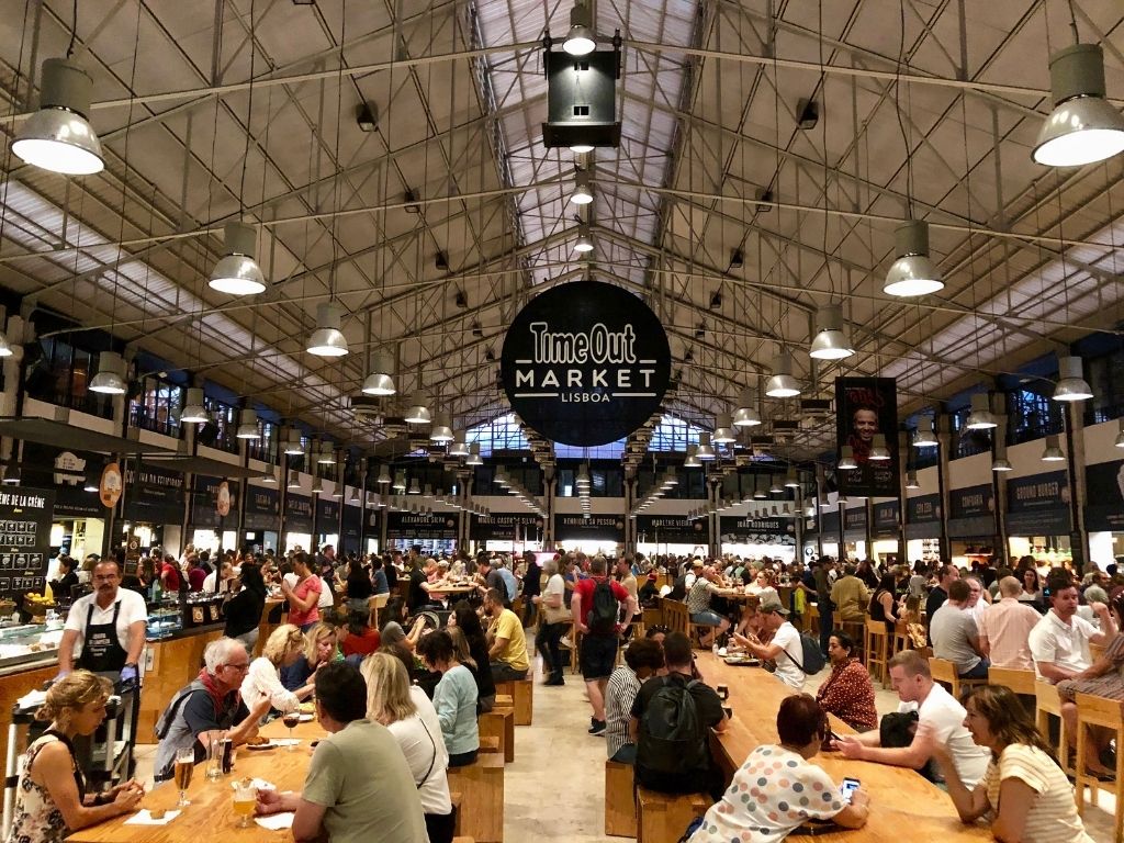 The time out market food hall in Lisbon. Hundreds of people sit at long tables under a black sign that reads "Time Out Market Lisboa"