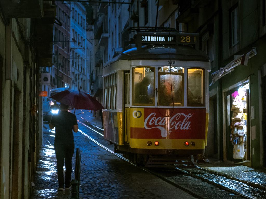 What to do in Lisbon when it rains? A yellow tram rolls down the street at night and a man walks past with an umbrella
